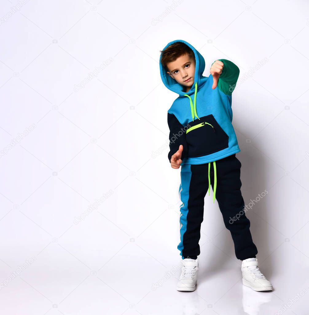 Cute stylish kid in colourful sportsuit standing with hood over head pointing fingers at camera. Full length portrait isolated on light grey