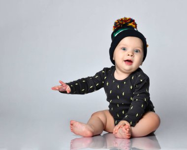 Siyah elbiseli, şapkalı, timsah izli, yalınayak bir bebek. Beyaz stüdyo arka planında izole bir şekilde yerde oturuyor. Kapat.