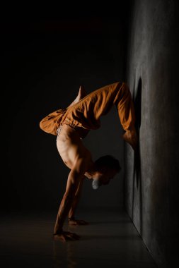 Kahverengi pantolonlu, çıplak gövdeli yaşlı yogi adam yoga yapıyor. Ellerinin üzerinde duruyor, tek ayak duvara yaslanıyor.