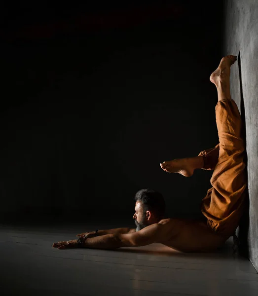 Elderly yogi male with naked torso, in brown pants is practicing yoga. Lying on the floor, leaning against wall by his legs