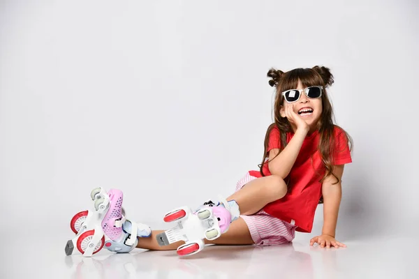 Hermosa chica posando sentada en el suelo, en una camiseta roja y pantalones cortos y un peinado de moda, en patines sobre sus pies — Foto de Stock