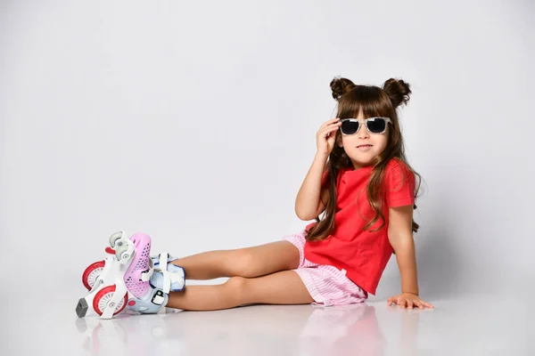 Hermosa chica posando sentada en el suelo, en una camiseta roja y pantalones cortos y un peinado de moda, en patines sobre sus pies — Foto de Stock