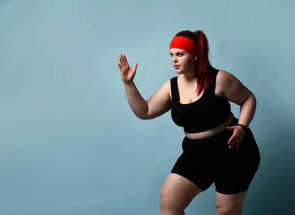 Serious concentrated young plus-size lady does exercises looking up with legs flexed and arms bent in elbows on blue background. Fitness. Copy space — Stock Photo, Image