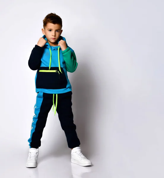 Şık pamuk jimnastik takımlı ciddi bir çocuk bacaklarını ayırıp ellerini ceplerinde tutuyor. Açık gri üzerinde izole edilmiş tam boy portre