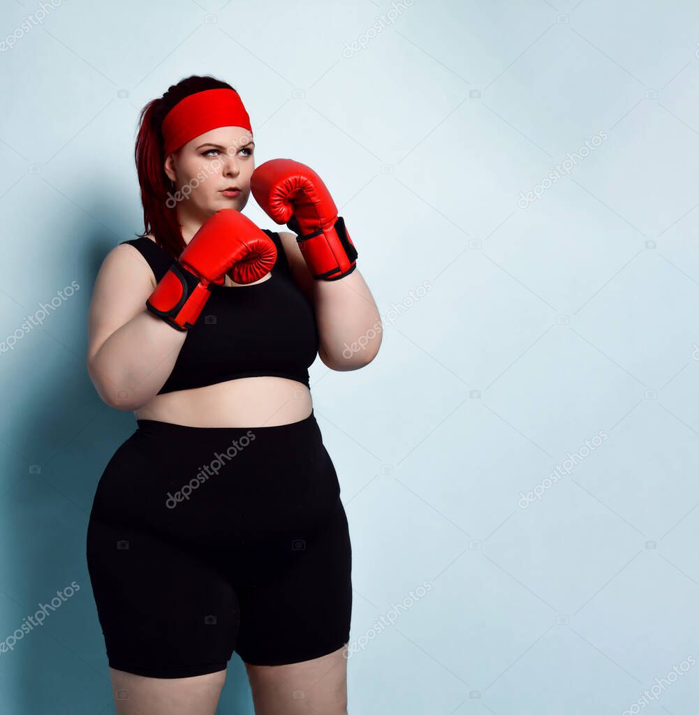 Thoughtful plump young girl in black sporty top, shorts and red boxing gloves looks up thinking over strategy in forthcoming fight. Copy space