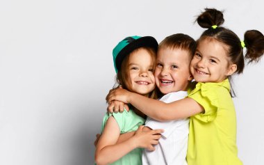 Studio portrait of children on a light background: full body shot of three children in bright clothes, two girls and one boy. Triplets, brother and sisters. hugging on camera. Family ties, friendship clipart