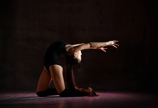 Молодая девушка гимнастка в черном спортивном теле и специальной обуви делает гимнастическую позу на темном фоне — стоковое фото