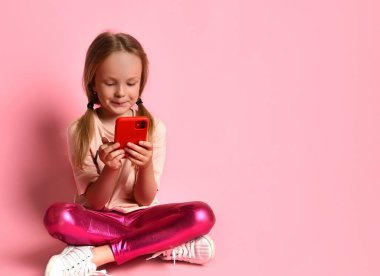 Tişörtlü, taytlı ve spor ayakkabılı küçük bir kız. Gülümsüyor, kırmızı akıllı telefonunu kullanıyor, pembe arka planda bacak bacak üstüne atmış bir şekilde yerde oturuyor. Kapat.