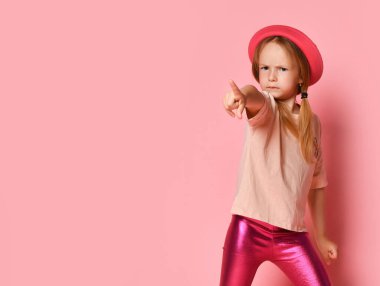 Şık bluzlu, taytlı ve Panama 'lı suratsız küçük kız kamerayı kızgın bir şekilde işaret ediyor. Kırpılmış çekim pembe üzerinde izole edildi, kopyalama alanı