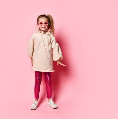Şık bir tayt ve kapüşonlu elbise içinde poz veren sarışın, neşeli bir kız. Stüdyoda pembe gözlüklerle poz veren mutlu genç çocuk. Parlak pozitif renkler.