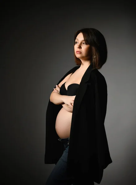 Mulher grávida em xale preto, calças e sutiã, barriga nua. Ela olhando para o lado, mãos dobradas, posando de lado no fundo cinza Imagem De Stock