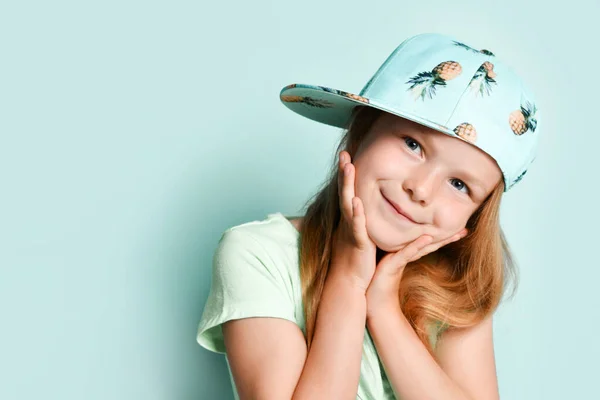 穿着T恤头戴凤梨帽的金发小子她面带微笑地抚摸着自己的脸,摆出青绿色的画室背景.靠近点 — 图库照片