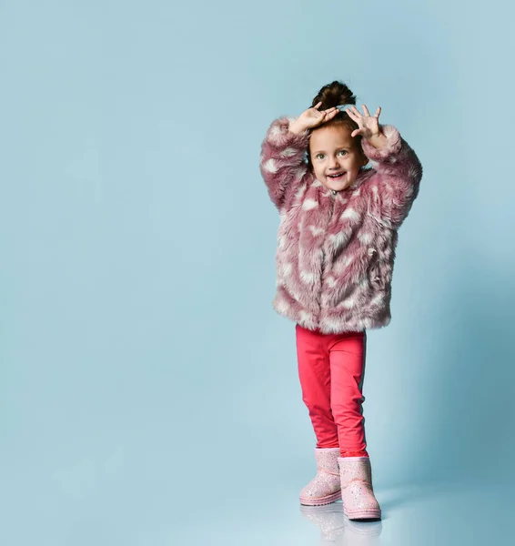 Маленька дівчинка, одягнена в рожеве хутро, штани та черевики. Вона посміхається, жестикулює, позує на синьому фоні. Повна довжина — стокове фото