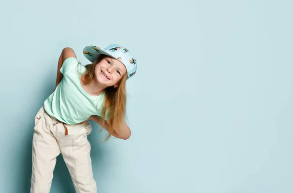Niño en camiseta, pantalones y gorra impresos con piñas. Se inclinó hacia adelante con los brazos separados, sonriendo, posando sobre fondo turquesa. De cerca. — Foto de Stock