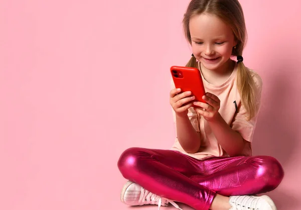 Niña con camiseta, polainas y zapatillas. Sonriendo, usando su teléfono inteligente rojo, sentado en el suelo con las piernas cruzadas contra el fondo rosa. De cerca. — Foto de Stock