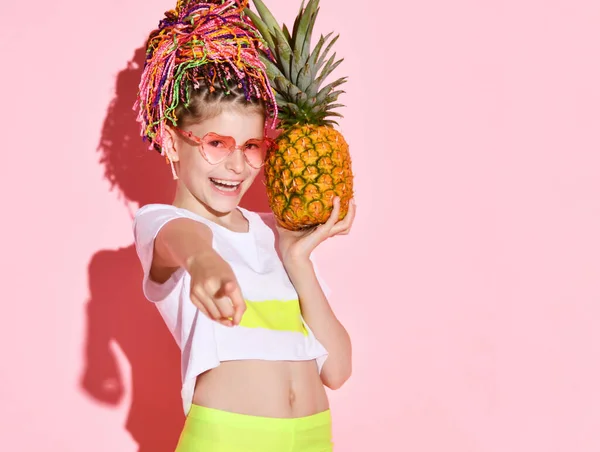 Active girl with rainbow colored african braids on head holding pineapple and pointing at camera with cheerful smile. Cropped shot isolated on pink — Stock Photo, Image