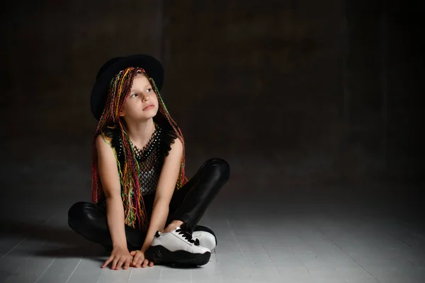 Adolescente com tranças africanas, em chapéu preto, perneiras, blusa de malha e tênis. Sentado no chão, posando no fundo escuro — Fotografia de Stock