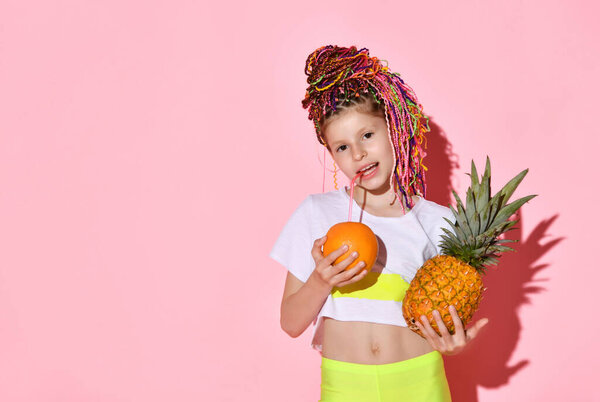 cute little girl with multi-colored pigtails on her head smiles happily, holding a pineapple and an orange in her hands. Cropped portrait isolated on pink, copy space. Childhood, emotions, summer.