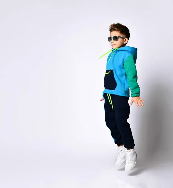 Little kid in sunglasses, colorful sport suit and sneakers. He performing a kick by his leg, posing isolated on white — Stock Photo, Image