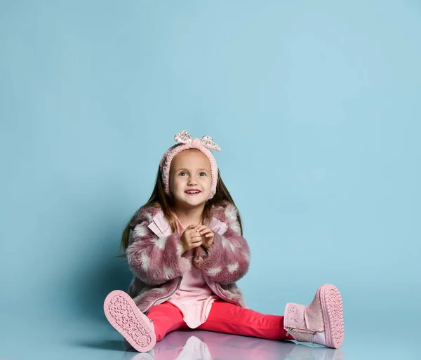 Kind in roze nepbontjas, broek en laarzen. Ze glimlacht, gebarentaal, zit op de vloer tegen een blauwe achtergrond. — Stockfoto