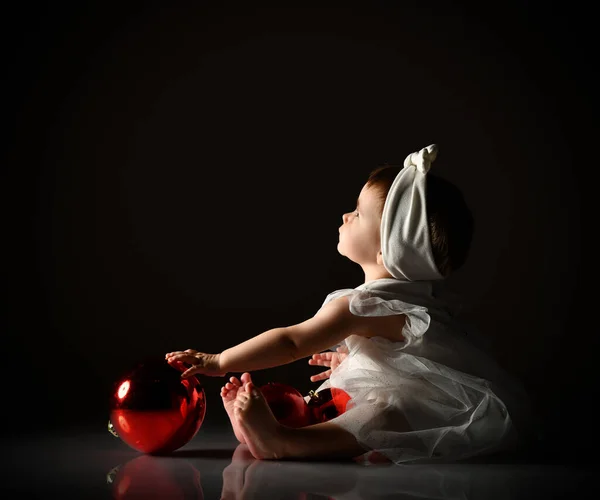白いヘッドバンドとドレス、裸足で赤ちゃんの女の子。2つの赤いボールを保持、上を見上げ、床に座っている。薄明色の背景. — ストック写真