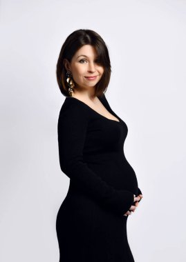 Dar elbiseli hamile kadın. Gülümsüyor, ellerini karnına koyuyor, yanlamasına beyaz poz veriyor.