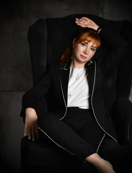 Une femme riche et autoritaire en costume de pantalon noir s'assoit dans une chaise haute dans une position confortable et vous regarde. Plan recadré avec fond gris foncé. — Photo