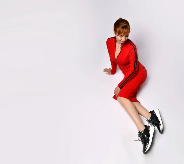 Draufsicht der schönen Frau in rotem Kleid mit schmalen Linien, die entspannt auf dem Boden sitzt und ihre schwarzen Turnschuhe betrachtet. Aufnahmen in voller Länge isoliert auf Weiß — Stockfoto
