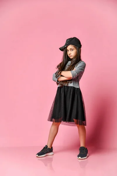 ピンクの背景に野球帽とスニーカーの暗いドレスの小さなヒップスターの女の子の肖像画 — ストック写真