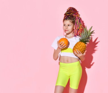 Elinde ananas tutarak portakaldan taze meyve suyu içen renkli afro demetli komik kız. Pembe üzerine üç çeyrek atış.