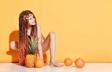 Afrikalı örgü saçlı, mayo giymiş genç bir kadın. Çömelip gülümsüyor, ananas ve portakallarla sarı arka planda poz veriyor.