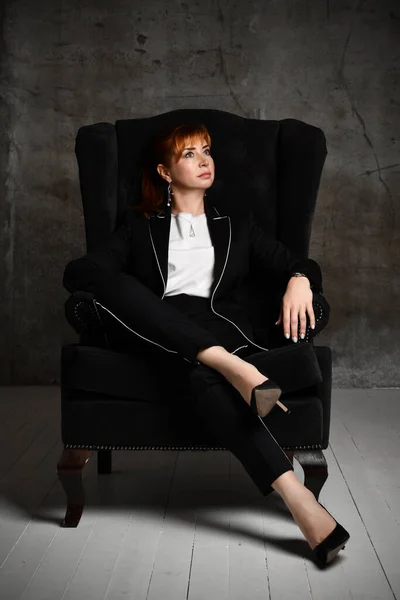 Nachdenkliche hübsche Frau in schwarzem Hosenanzug und Stöckelschuhen sitzt im Sessel im dunklen Raum und schaut nachdenklich zur Seite — Stockfoto