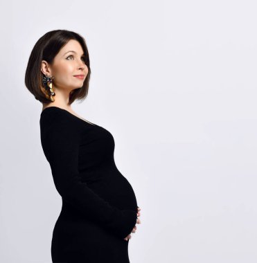 Siyah dar elbiseli, kocaman küpeli esmer kız. Gülümsüyor, elini karnına koyuyor, yanlamasına bembeyaz poz veriyor.