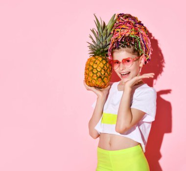 Gökkuşağı renkli Afrika örgülü aktif bir kız kafasında ananas tutuyor ve neşeli bir gülümsemeyle kameraya bakıyor. Kesilmiş atış pembeye izole edilmiş.