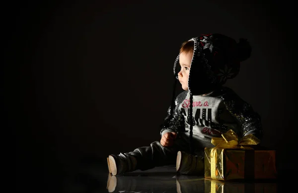 Klein kind met grijze hoed, pak en laarzen. Ze kijkt omhoog, houdt gouden geschenkdoos vast, zit op de vloer. Zwarte achtergrond. Sluiten. — Stockfoto