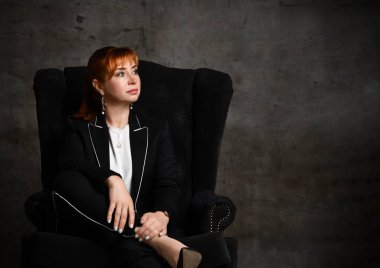 Siyah takım elbiseli, kendine güvenen kızıl saçlı bir kadın koltukta rahat bir şekilde oturuyor ve bir bacağı diğerinin üzerinde. Koyu gri arkaplanda kırpılmış çekim
