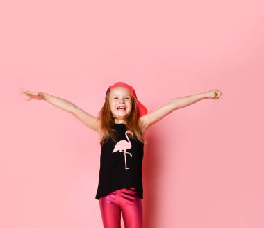 Şapkalı küçük bir kız, baskılı flamingo ve taytlı siyah bir bluz. Ellerini aralayıp gülümsüyor, pembe arka planda poz veriyor.