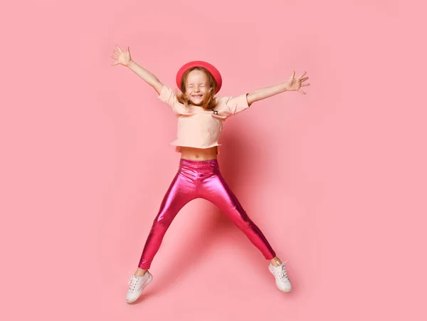 Kleine vrouw met hoed, t-shirt, legging en witte sneakers. Ze glimlacht en springt op met gesloten ogen. Poseren op een roze achtergrond. Totale lengte — Stockfoto