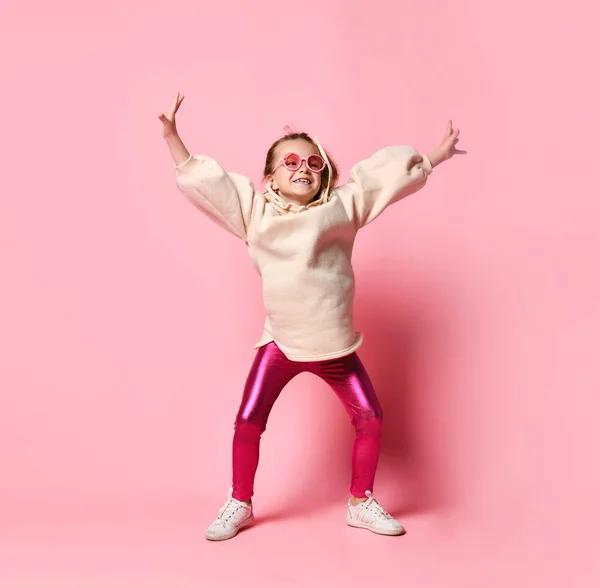 Chica alegre rubia posando en un traje elegante de polainas y sudaderas con capucha. Feliz adolescente posando en un estudio con gafas rosas. Colores positivos brillantes. — Foto de Stock