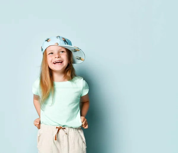 Радісна маленька дівчинка в синьому літньому вбранні і стильна шапка з ананасовим візерунком голосно сміється, тримаючи руки на талії. Ізольовано на синьому, простір копіювання — стокове фото