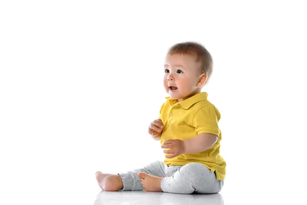 Pequeño bebé en un polo amarillo se sienta en el suelo, se ríe, toca sus piernas y mira a un lado en la esquina superior sobre un fondo blanco. — Foto de Stock