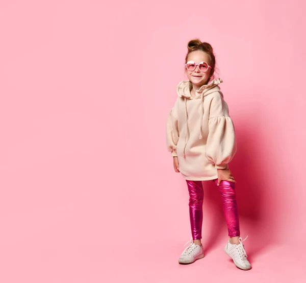Маленька дівчинка в стильному рожевому одязі і круглих окулярах позує спираючись вперед. Повнометражний знімок ізольовано на рожевому, простір копіювання . — стокове фото