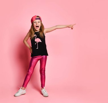 Kendine güvenen, beyzbol şapkası takmış, eli kalçasında poz veren küçük bir kız. Tam uzunlukta çekim pembe üzerinde izole edildi, kopyala boşluk