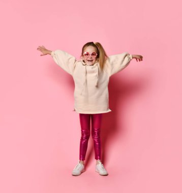 Gözlüklü, şık kapüşonlu, taytlı ve spor ayakkabılı küçük sarışın çocuk. Ellerini açıyor ve gülümsüyor, pembe arka planda poz veriyor. Tam uzunluk
