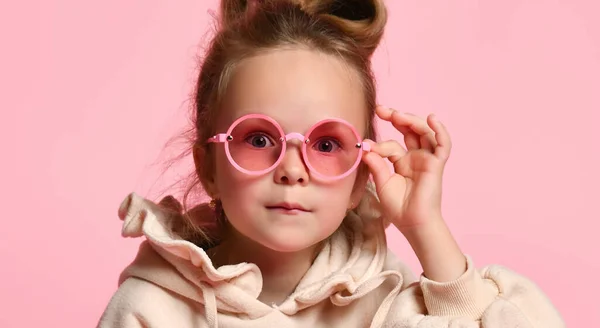 Portret van een blondje in een bril, een stijlvolle hoodie. Jou aandachtig aankijken, poserend op een roze studio achtergrond. Jeugd, oprechte emoties, mode, reclame. — Stockfoto