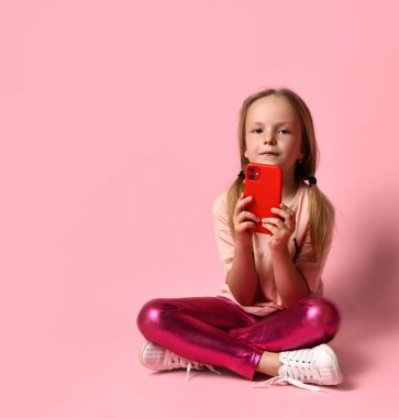 Tişörtlü, taytlı ve spor ayakkabılı küçük bir kız. Gülümsüyor, kırmızı akıllı telefonunu kullanıyor, pembe arka planda bacak bacak üstüne atmış bir şekilde yerde oturuyor. Kapat.