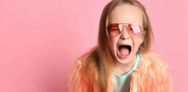 Blond kind met zonnebril, turquoise t-shirt en nepbontjas. Ze schreeuwt luid terwijl ze poseert tegen een roze studio achtergrond. Sluiten. — Stockfoto