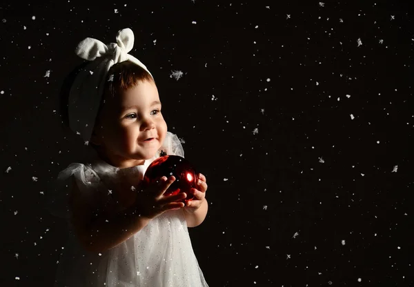 Малыш в белой повязке и платье, босиком. Она позирует с двумя красными шариками. Сумерки, черный фон. Новый год, праздники. — стоковое фото