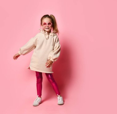 Şık bir tayt ve kapüşonlu elbise içinde poz veren sarışın, neşeli bir kız. Stüdyoda pembe gözlüklerle poz veren mutlu genç çocuk. Parlak pozitif renkler.