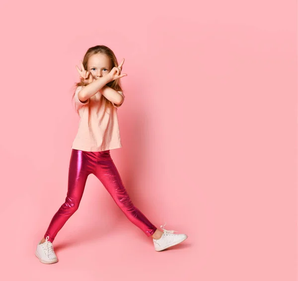 Симпатична маленька дівчинка стоїть з ногами в ширину і ховає обличчя за двома схрещеними руками. Повнометражний знімок ізольовано на рожевому, простір копіювання — стокове фото